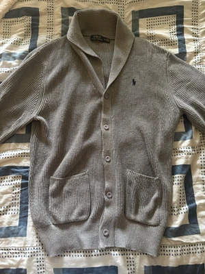 Grå kofta från Polo Ralph Lauren - Snygg grå stickad kofta från Polo Ralph Lauren med sjalkrage, knäppning framtill och två stora fickor. Klassisk broderad logga på bröstet. Perfekt för lager på lager och chill stil.