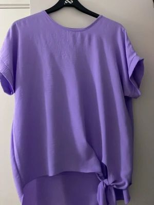 Ljuslila topp med knyt i sidan - Säljer en ljuslila topp med korta ärmar och rund halsringning. Toppen har en snygg knytdetalj i sidan och är lite längre baktill för en avslappnad look. Perfekt att styla med jeans eller kjol.