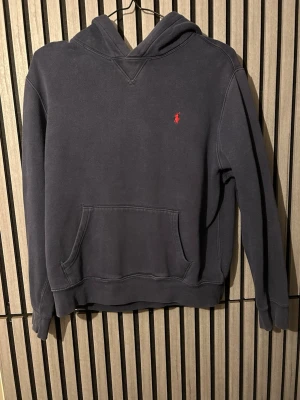Mörkblå hoodie med röd logga - Mörkblå hoodie med huva och känguruficka framtill. Inga håll vad jag kan se använd rätt mycket.