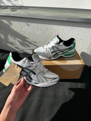 Asics gel kayano 14 malachite green - Snygga sneakers från Asics i vitt, silver och grönt med svarta detaljer. Skorna har en sportig design med mesh och syntetmaterial, samt en bekväm sula som ger bra stöd. Perfekta för dig som gillar streetwear och vill ha en fräsch look.