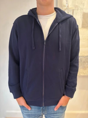Mörkblå hoodie med dragkedja - Mörkblå hoodie med hel dragkedja, två fickor framtill och justerbar huva med snören. Tillverkad i mjukt tyg och har ribbade muddar vid ärmar och nederkant. Enkel och clean design som passar till jeans och t-shirt.