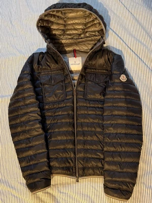 Moncler Clovis down jacket - Tja, säljer denna svarta Moncler Clovis dunjackan. Jackan har en avtagbar luva och är i väldigt fint skick. Extra orginalknappar medföljer. Storlek 4, passar M/L. Modellen på bilden är 183cm. Kvitto saknas men jackan är självklart äkta. Kontakta mig privat vid fler frågor eller funderingar! 