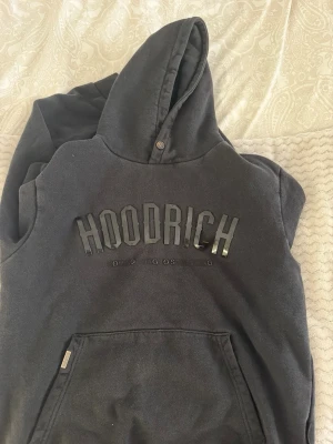 Svart hoodie från Hoodrich med tryck - Säljer en svart hoodie från Hoodrich med stor logga i blankt tryck på bröstet. Klassisk känguruficka framtill och huva med knappdetalj. Mjuk insida och avslappnad passform, perfekt för en streetwear-look.