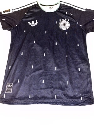 Svart Adidas fotbollströja Tyskland - Säljer en svart fotbollströja från Adidas med Tysklands landslagslogga på bröstet. Tröjan har vita detaljer, diskret mönster och klassiska Adidas-ränder på axlarna. Tillverkad i lätt och ventilerande material med Climacool-teknologi.