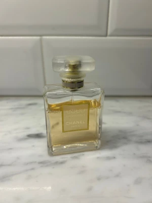 Coco Mademoiselle parfym från Chanel - Elegant parfymflaska från Chanel, Coco Mademoiselle. Flaskan är rektangulär i genomskinligt glas med en frostad, fyrkantig kork och guldfärgade detaljer. Innehållet är ljus bärnstensfärgat och etiketten är stilrent guldig med vit text.