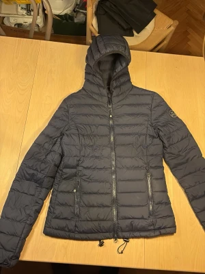 Mörkblå dunjacka från North Sails - Snygg mörkblå dunjacka från North Sails med huva och quiltad design. Yttermaterial i nylon och elastan, fodrad med polyester och fylld med 90% dun och 10% fjädrar. Perfekt för kalla vinterdagar och har praktiska innerfickor.