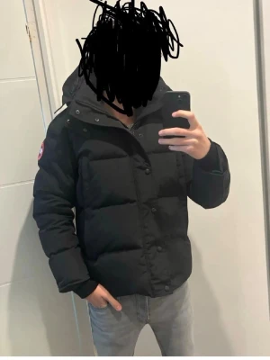 Svart dunjacka med pälskrage - Säljer en svart dunjacka med avtagbar pälskrage och huva. Jackan har en patch på ärmen, dragkedja och knappar framtill samt flera fickor. Perfekt för kalla vinterdagar och riktigt varm. Klassisk, stilren modell som passar till det mesta.