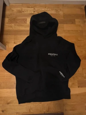 Svart Essentials Fear of God hoodie - Säljer en svart hoodie från Essentials Fear of God med vit logga på bröstet och på armen. 