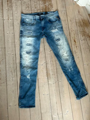 Blå slitna jeans från Replay - Säljer ett par blå jeans från Replay med slitningar och trasiga detaljer på både fram- och baksida. Jeansen har fem fickor, normal midja och smal passform. Perfekta för dig som gillar en trendig och avslappnad stil.