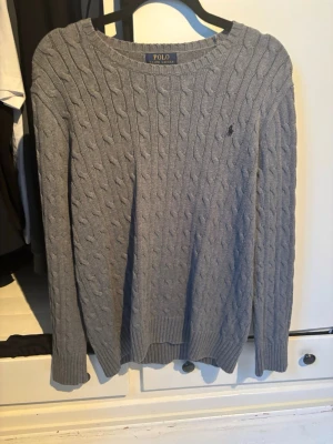 Grå kabelstickad tröja från Polo - Snygg grå kabelstickad tröja från Polo Ralph Lauren med rund halsringning och diskret logga broderad på bröstet. Passar S och M 