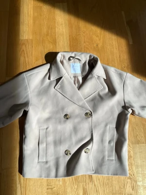 Beige skepparkavaj med dubbelknäppning - Knapparna går även att öppna! Stilren beige skepparkavaj med klassisk dubbelknäppning och bred krage. Jackan har två raka fickor framtill och är tillverkad i ett mjukt tyg med slät yta. Perfekt för dig som gillar minimalistisk och tidlös stil.