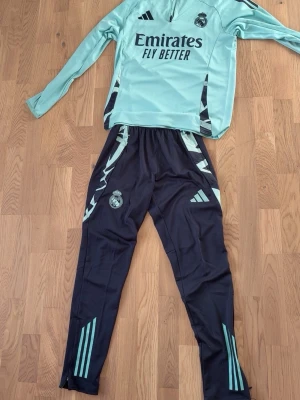 Real Madrid träningsset Adidas - Snyggt träningsset från Real Madrid och Adidas. Överdelen är ljusgrön med svarta detaljer och dragkedja vid halsen, medan byxorna är svarta med ljusgröna ränder och klubbmärke. Perfekt för fotbollsträning eller chill. Materialet är mjukt och stretchigt funktionsmaterial.