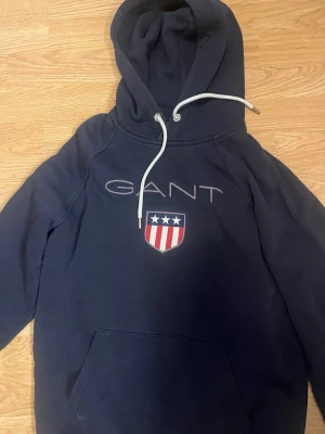 Mörkblå hoodie från GANT med logga - Snygg mörkblå hoodie från GANT med stor logga och sköld med stjärnor och ränder på bröstet. Klassisk känguruficka framtill och vita dragsnören i huvan. Perfekt för en avslappnad och sportig stil. Stolek M men är liten i storleken så passar S bättre