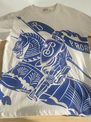 Vit burbbery t-shirt med blått riddarmotiv - Snygg vit t-shirt med stort blått tryck av en riddare på häst och texten 'PRORL'. T-shirten har rund hals och korta ärmar. Perfekt för dig som gillar unika och grafiska prints.