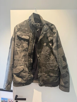 Cammo jacka  - Snygg vindjacka i grått camouflage-mönster med flera praktiska fickor och hög krage. Köptes för några månader sen i en vintage butik i Prag. 