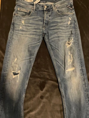 Replay jeans  - Säljer ett par blå jeans med slitningar och hål på både fram- och baksida. Jeansen har klassisk femficksmodell, knappgylf och ljus tvätt med tydliga slitdetaljer. Perfekta för en avslappnad och trendig look.