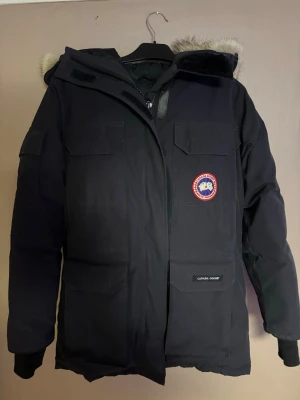 Svart parkas från Canada Goose - ⚠️Den är 100% äkta!⚠️Snygg svart parkas från Canada Goose med klassisk patch på bröstet. Jackan har flera stora fickor, huva med pälskant och robust dragkedja. Perfekt för kalla dagar och riktigt varm. Materialet är slitstarkt och vindtätt. Storlek S men skulle säga att den motsvarar en storlek M.