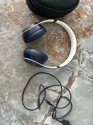Blå och vita over-ear hörlurar - Snygga over-ear hörlurar i mörkblått och vitt med vadderade kåpor och justerbart huvudband. Hörlurarna är hopfällbara och levereras med en svart laddkabel och ett smidigt fodral. Perfekta för musik, gaming eller videos.