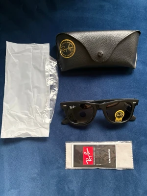 Svarta Ray-Ban Wayfarer solglasögon - Klassiska svarta Ray-Ban Wayfarer solglasögon med tidlös design och mörka glas. Modellen är handgjord i Italien. Kommer med originalfodral och putsduk. Endast provade! Legat undan i låda utan att användas. Litet paket så frakten är inte alls dyr!