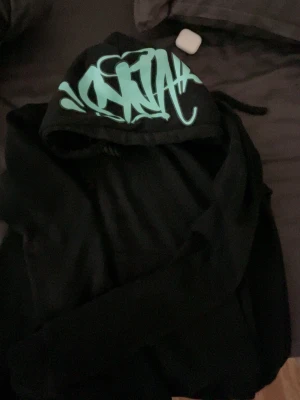 Svart hoodie från syna world - Säljer en svart hoodie från syna world  med ett fett turkost graffiti-tryck på huvan. Klassisk passform med huva och dragsnören. Perfekt för dig som gillar streetwear och vill sticka ut lite extra. Den är storlek m men passar mer som s