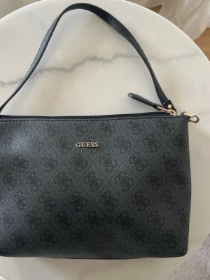 Svart liten väska från Gyess - Säljer en svart liten väska från Guess. Aldrig använd så som nyskick. Väskan har dragkedja och fint mönster💕