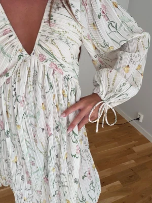 Blommig vit kortklänning med knyt - Söt vit kortklänning med blommigt mönster i pastellfärger. Klänningen har v-ringning, långa ärmar med knyt vid ärmslut och volangdetaljer. Tillverkad i ett lätt, plisserat material som passar perfekt för sommarens varma dagar.