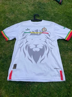 Senegal fotbollströja Puma vit - Säljer en vit Senegal fotbollströja från Puma med stort lejonmotiv på framsidan. Tröjan har gröna, gula och röda detaljer på ärmslut och bröstet samt Senegals landslagsemblem. Tillverkad i lätt och ventilerande material, perfekt för träning eller match.