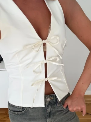 Vit väst med knyt och v-ringning - Säljer en snygg vit väst med djup v-ringning och tre dekorativa knyt framtill. Västen har en croppad passform och är tillverkad i ett slätt tyg som ger en stilren look. Perfekt att styla över en topp eller direkt mot huden för en trendig outfit.
