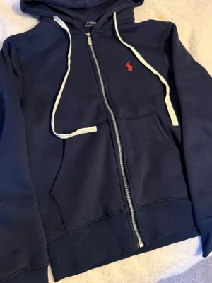 Mörkblå hoodie från Polo Ralph Lauren - Mörkblå hoodie med dragkedja från Polo Ralph Lauren. Har vit dragsko i huvan, kängurufickor och det klassiska röda Polo-logot broderat på bröstet. Tillverkad i mjukt sweatshirtmaterial, perfekt för chill dagar.