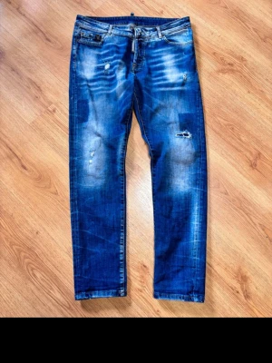 Blå slitna jeans från Dsquared2 - Säljer ett par blå jeans från Dsquared2 i modell slim fit. Jeansen har slitningar och distressed detaljer på benen samt klassisk femficksdesign. Tillverkade i Italien och har snygga kontrastsömmar. Perfekta för en trendig och avslappnad stil. Midja 45cm längd 101cm