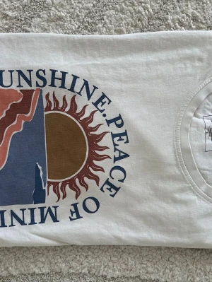 Vit t-shirt med soltryck och text - Vit t-shirt med ett stort tryck framtill som visar en sol, berg och texten 'SUNSHINE. PEACE OF MIND' i blått. T-shirten har rund hals och korta ärmar. Perfekt för en chill och avslappnad stil.