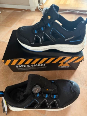 Svarta sneakers Monitor BOA-system - Skyddsskor från Monitor, modell Paradox R monitor. Skorna är svarta med blå detaljer och har ett BOA-snörningssystem. De är designade för att vara säkra och smarta, med fokus på lätthet, andningsförmåga och hållbarhet för långa arbetsdagar. Skorna är märkta med ESD.  Nya med lådan