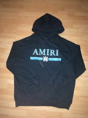 Svart Amiri hoodie    - Säljer en svart hoodie från Amiri med stor vit logga och blått tryck på bröstet. Klassisk huva och känguruficka framtill. Hoodien har ribbade muddar vid ärmslut och nederkant. Perfekt för dig som gillar streetwear och vill sticka ut.