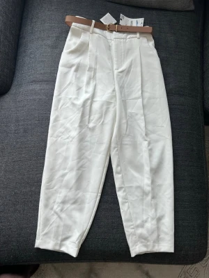 Vita kostym byxor från Zara med brunt bälte - Stilrena klassiska vita kostymbyxor från Zara med hög midja och pressveck framtill. Byxorna har två fickor bak och kommer med ett snyggt brunt bälte. Perfekta för en fräsch och avslappnad look.