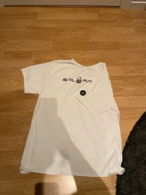 Vit Sail Racing t-shirt med tryck - Säljer en vit t-shirt från Sail Racing med svart logotyptryck på bröstet och en rund svart detalj på ärmen. T-shirten har klassisk passform och korta ärmar, perfekt till vardagslooken. Materialet känns mjukt och skönt mot huden.