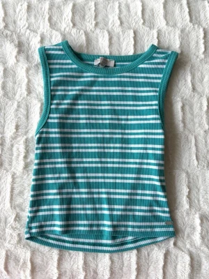 Randig turkos ribbad linnetopp - Snygg ärmlös linnetopp i turkos och vitt med horisontella ränder. Toppen är ribbad och har en rund halsringning. Perfekt för varma dagar och enkel att matcha med jeans eller shorts.