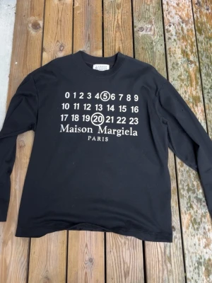 Svart långärmad tröja Maison Margiela - Cool svart långärmad topp från Maison Margiela med ikoniskt tryck framtill i vitt. Rund halsringning och relaxed passform. Perfekt för dig som gillar streetstyle och vill ha något unikt i garderoben.