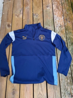 Blå Manchester City träningströja Puma - Snygg blå långärmad träningströja från Puma med Manchester City-logga på bröstet. Tröjan har halv dragkedja, vita och ljusblå detaljer på axlar och sidor samt guldiga Puma-detaljer. Perfekt för fotbollsträning eller sportiga dagar.