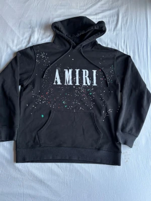 Amiri hoodie  - Säljer en svart hoodie från Amiri med stor vit logga framtill och färgstänk i vitt, grönt och rött. Hoodien har huva med dragsko, magficka och ribbade muddar vid ärmar och nederkant. Perfekt för dig som gillar streetwear och vill sticka ut.