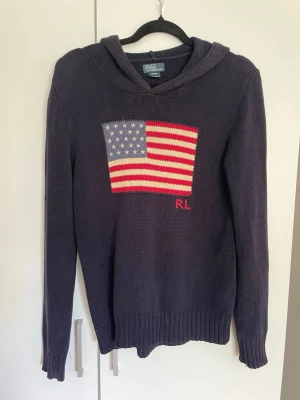 Marinblå Ralph Lauren hoddie - Stickad hoodie i marinblå färg med amerikansk flagga framtill och RL-broderi. Tröjan har långa ärmar, ribbade muddar och huva. Perfekt för dig som vill ha en casual look. Storlek 14-16 barn, passar om man är ca 160-170 lång