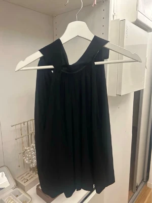 Svart kort klänning med knyt - Snygg svart kort klänning med knyt vid axeln. Klänningen har en lös passform och är ärmlös, vilket ger en stilren och modern look. Perfekt för dig som gillar minimalistisk stil och vill ha något enkelt men ändå trendigt.