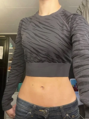 Mörkgrå croppad zebramönstrad topp - Säljer en croppad långärmad topp i mörkgrått med zebramönster. Toppen har bred mudd i midjan och är figurnära med rund halsringning. Perfekt för dig som gillar trendiga och unika plagg. Funkar väldigt bra för träning då den är luftig och sval