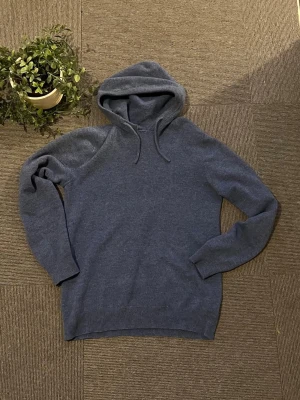 Mörkblå hoodie med snörning - En stilren mörkblå merinoull hoodie med snörning i huvan. Hoodien har en mjuk, stickad känsla och ribbade muddar vid ärmslut och nederkant. Perfekt för dig som gillar en enkel och clean look.