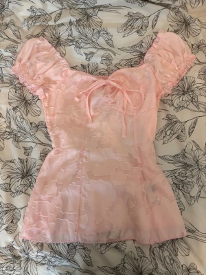 Ljusrosa blus med puffärm och rosett - Supersöt ljusrosa blus med puffärmar och en liten rosett framtill. Blusen har ett subtilt blommigt mönster i tyget.