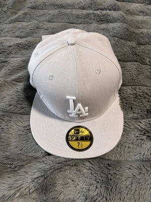 Vit LA 59FIFTY keps - Snygg vit 59FIFTY keps från New Era med LA-logga broderad framtill. Klassisk flat brim och strukturerad passform. Kepsen är gjord i ett tåligt tyg och har officiella MLB-detaljer på insidan. Perfekt för dig som gillar streetwear och sportig stil. Priset är inte hugget i sten och det finns 2st ifall ni vill köpa en till