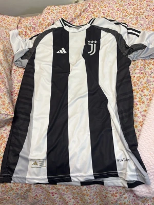Juventus matchtröja svartvit - Juventus officiella matchtröja från Adidas i svartvitt randigt med klubbmärke och tre stjärnor på bröstet. Tröjan har VLAHOVIC och nummer 9 på ryggen samt detaljer som HEAT.RDY-tryck och Serie A-märke på ärmen. Tillverkad i funktionsmaterial för sport.