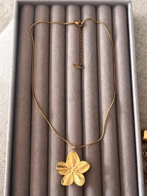 Guldigt halsband med blomma - Snyggt halsband i guldfärg med en stor, detaljrik blomma som hänge. Kedjan är tunn och har ett justerbart lås för perfekt passform. Perfekt accessoar för att lyfta vilken outfit som helst med en elegant touch.