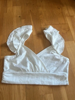 Vit omlottopp med volang från SHEIN - Söt vit omlottopp från SHEIN med broderade detaljer och volangprydda axelband. Toppen är ärmlös och har en feminin v-ringning. Perfekt för sommarens alla tillfällen och ger en romantisk vibe till din outfit.