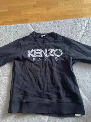 Svart sweatshirt från Kenzo Paris - Säljer en svart sweatshirt från Kenzo Paris med stort vitt tryck på bröstet. Tröjan har rund halsringning, ribbade muddar vid ärmslut och nederkant samt en relaxed fit. Perfekt för dig som gillar streetwear och vill ha något stilrent men ändå med en twist.