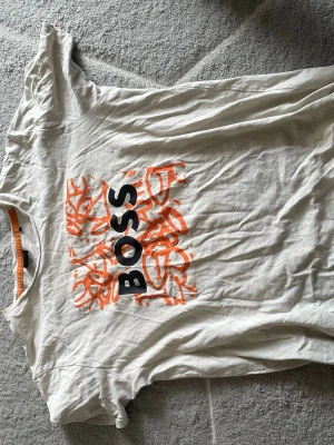 Vit BOSS t-shirt med orange tryck - Snygg vit t-shirt från BOSS med ett stort grafiskt tryck i orange och svart text på bröstet. Klassisk rund halsringning och korta ärmar. Perfekt för dig som gillar streetstyle och vill ha en enkel men ändå unik look.